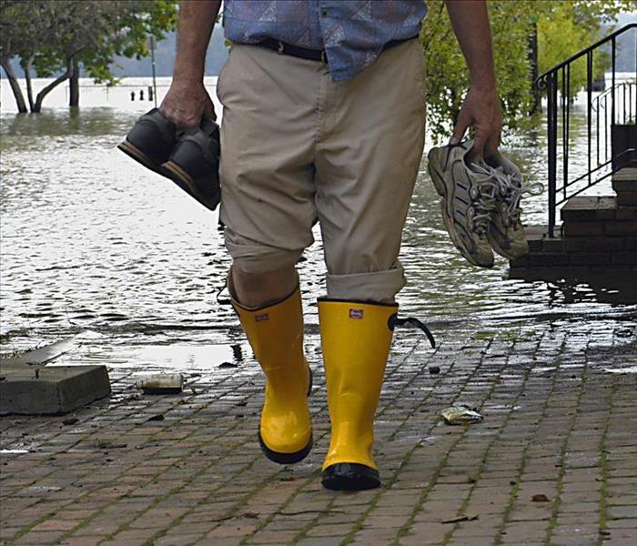 Image of a person walking on the street while holding shoes, behind him we can see a flooded street. 